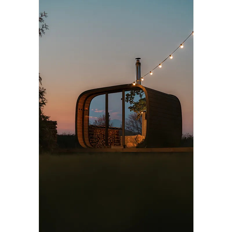 Leil Round Cube Single – Premium Gartensauna 1 Raum von Leil Saunas im Garten bei Sonnenuntergang mit Holzlagerung und Lichterkette