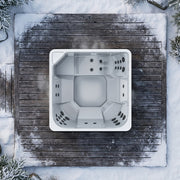 Draufsicht auf den weißen Novitek L-1 Outdoor Whirlpool mit quadratischem Design, eingelassen in verschneite Holzterrasse, umgeben von Schnee und Nadelbäumen