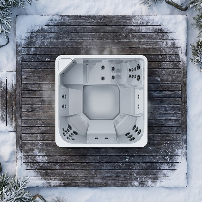 Draufsicht auf den weißen Novitek L-1 Outdoor Whirlpool mit quadratischem Design, eingelassen in verschneite Holzterrasse, umgeben von Schnee und Nadelbäumen