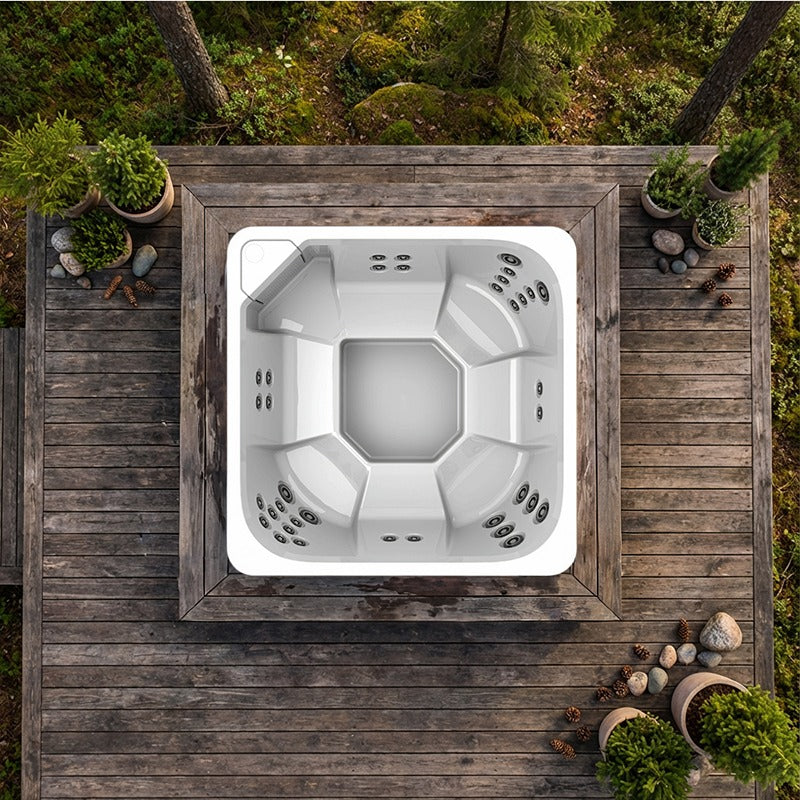 Vogelperspektive eines weißen quadratischen Novitek M-0 Outdoor Whirlpools mit Massagedüsen auf Holzterrasse, umgeben von grünen Pflanzen und Natursteinen