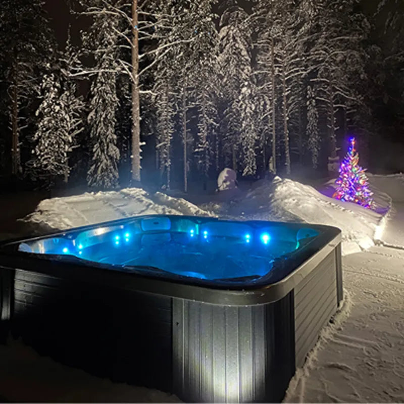 Novitek L-0 Outdoor Whirlpool mit LED-Beleuchtung in verschneiter Winterlandschaft mit Wald und leuchtendem Weihnachtsbaum