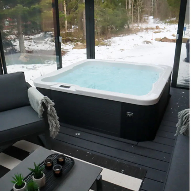 Novitek L-0 Outdoor Whirlpool mit türkisblauem Wasser auf schwarzem Untergestell, umgeben von großen Glasfenstern mit Blick auf schneebedeckte Waldlandschaft