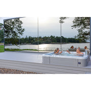 Moderner Novitek L-1 Whirlpool mit weißem Rahmen auf Holzterrasse, mehrere Personen entspannen im sprudelnden Wasser, große Glaspaneele bieten Blick auf See und Waldlandschaft.
