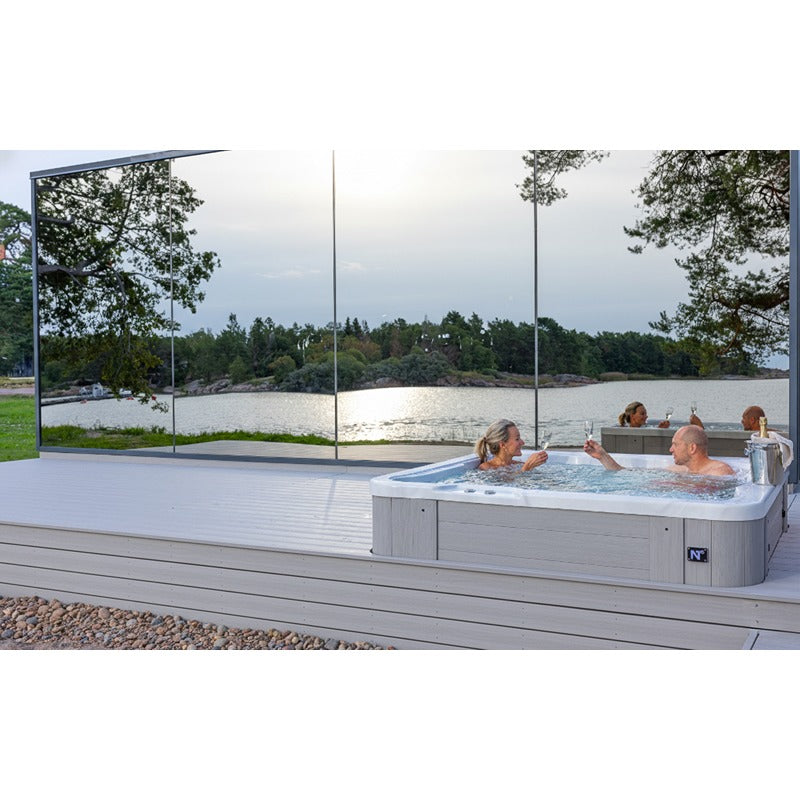 Moderner Novitek L-1 Whirlpool mit weißem Rahmen auf Holzterrasse, mehrere Personen entspannen im sprudelnden Wasser, große Glaspaneele bieten Blick auf See und Waldlandschaft.