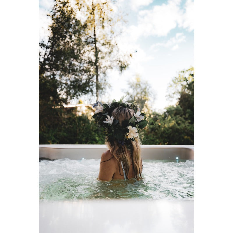 Frau mit Blumenkranz badet in Novitek S-0 Outdoor Whirlpool umgeben von Natur und Bäumen bei Tageslicht