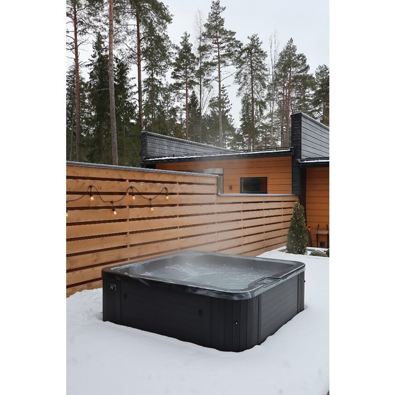 Moderner schwarzer Whirlpool im Schnee vor einer Holzfassade mit Metallklimaanlagen und Kiefernwald im Hintergrund