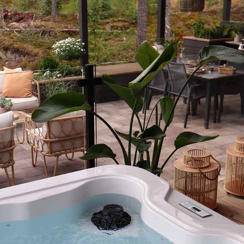 Novitek S-0 Outdoor Whirlpool mit blauem Wasser in sonniger Gartenlandschaft, umgeben von Loungemöbeln und grünen Pflanzen