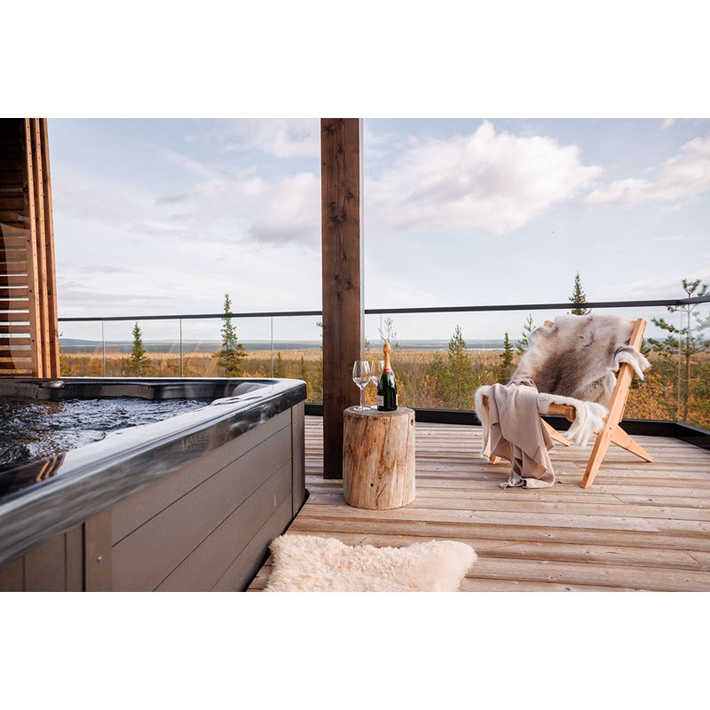 Novitek S-2 Outdoor Whirlpool auf einer Holzterrasse mit Panoramablick auf Waldlandschaft, daneben Liegestuhl mit Decke und Weinglas auf Holzhocker