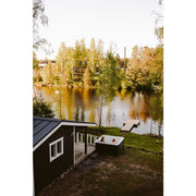 Novitek L-2 Outdoor Whirlpool am See gelegen, zwei Personen entspannen im weißen Whirlbecken vor schwarzem Gartenhaus mit Blick auf herbstliche Wald- und Wasserlandschaft.