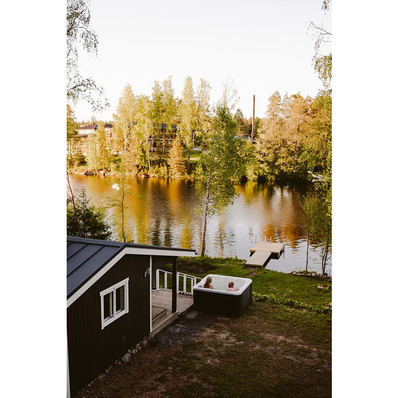 Novitek L-2 Outdoor Whirlpool am See gelegen, zwei Personen entspannen im weißen Whirlbecken vor schwarzem Gartenhaus mit Blick auf herbstliche Wald- und Wasserlandschaft.