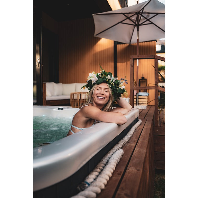 Frau in Blumenkranz genießt abends den Novitek S-1 Outdoor Whirlpool mit sprudelndem Wasser, umgeben von grauem Sonnenschirm und eleganter Terrasse