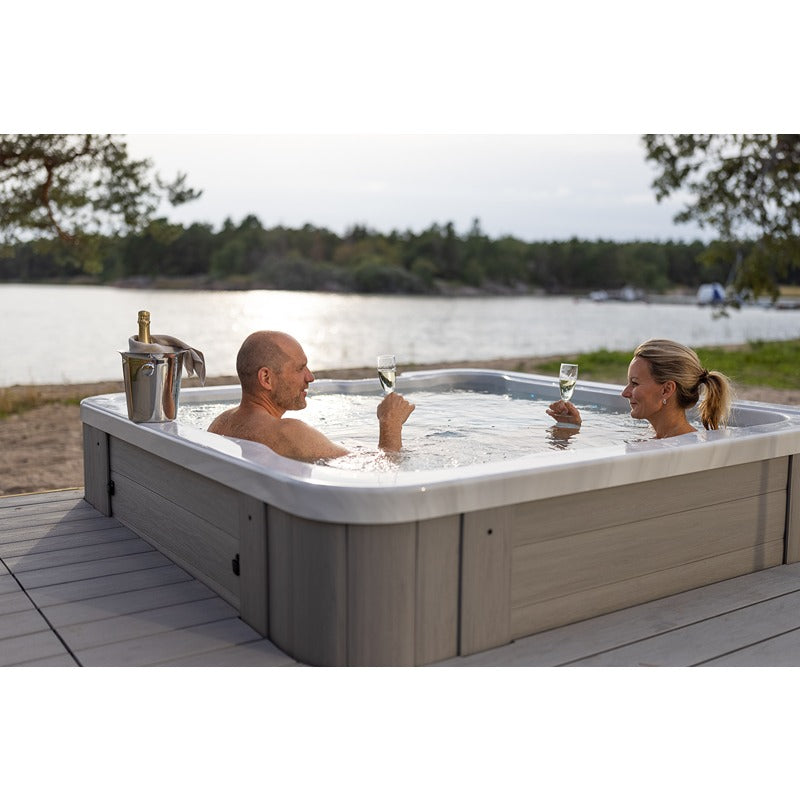 Paar genießt Whirlpool mit Seeblick, Champagner und Gläser auf Holzterrasse, Wald im Hintergrund