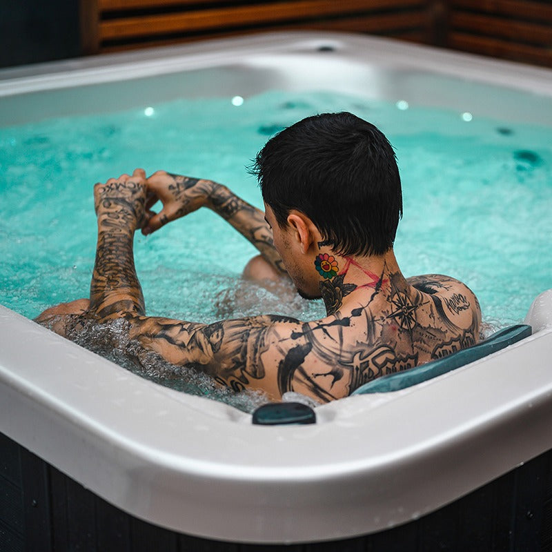 Mann mit Tätowierungen entspannt sich im türkisblauen Novitek S-1 Outdoor Whirlpool mit sprudelndem Wasser und weißem Rahmen