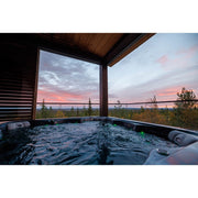 Novitek M-1 Outdoor Whirlpool mit sprudelndem Wasser auf einer Terrasse mit Panoramablick auf See und Wald bei Sonnenuntergang