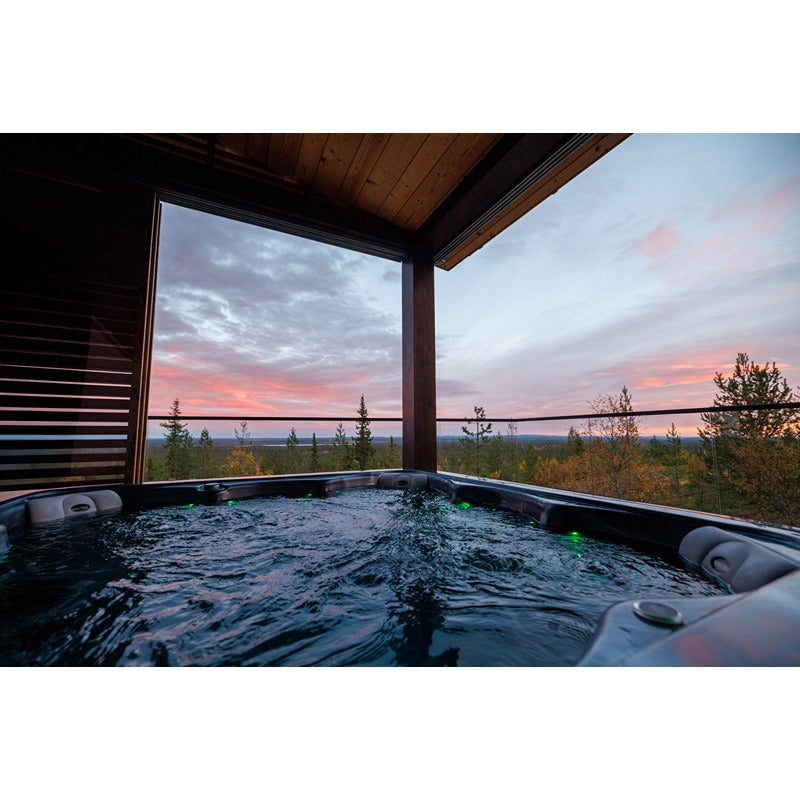 Novitek M-1 Outdoor Whirlpool mit sprudelndem Wasser auf einer Terrasse mit Panoramablick auf See und Wald bei Sonnenuntergang