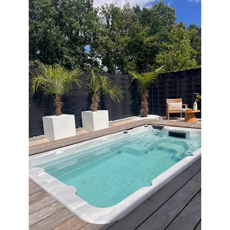 Passion Spas Aquatic 1 Eco Swim Spa für 3 Personen in modernem Garten mit Holzterrasse, Palmen und schwarzem Zaun