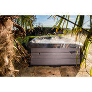 Platinum Spas Maximus Outdoor Whirlpool mit sprudelndem Wasser auf einer Terrasse, umrahmt von grünen Pflanzen und Palmen, in sonniger Gartenumgebung.