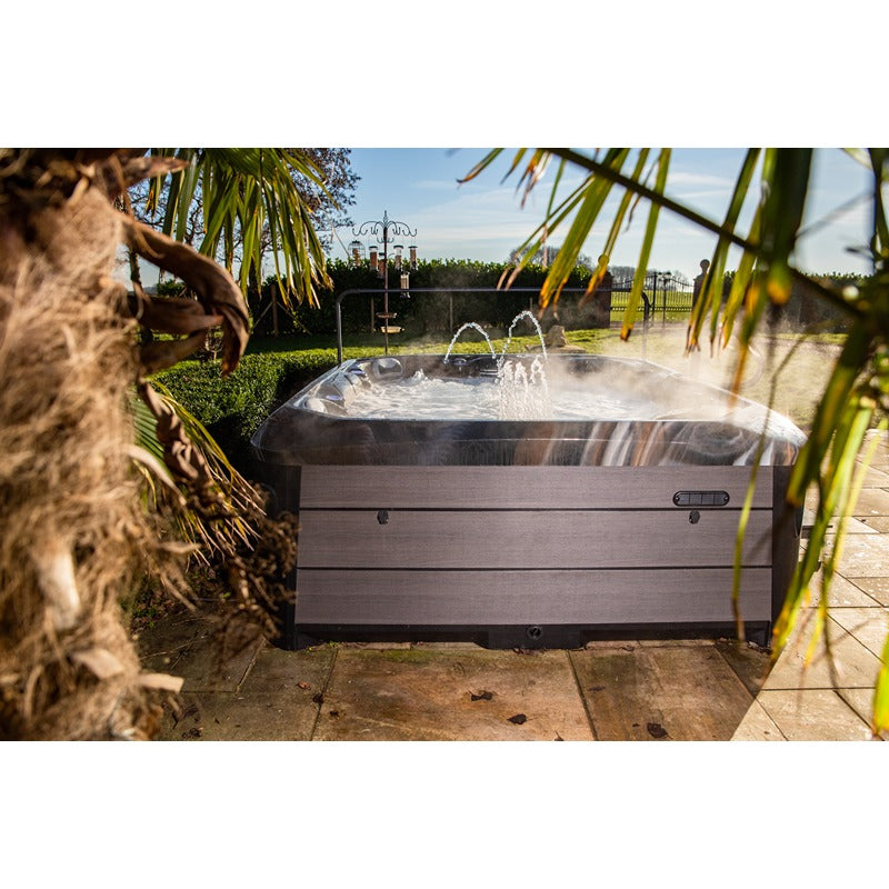 Platinum Spas Maximus Outdoor Whirlpool mit sprudelndem Wasser auf einer Terrasse, umrahmt von grünen Pflanzen und Palmen, in sonniger Gartenumgebung.