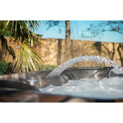 Platinum Spas Maximus Outdoor Whirlpool mit sprudelndem Wasser, Wasserfontäne und Entspannungsbereich unter freiem Himmel, umgeben von tropischen Pflanzen und Gartenmauer.