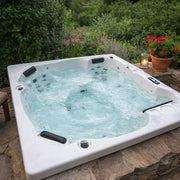Portcril Emotion 5 Whirlpool für 5 Personen mit sprudelndem Wasser, eingebaut in eine Naturstein-Terrasse, umgeben von blühendem Garten mit Kerzenlaterne.