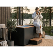 Frau im weißen Bademantel nutzt Swedish Cold Generation 4S Eisbad mit schwarzem Design auf einer Holzterrasse mit Seeblick und Waldkulisse