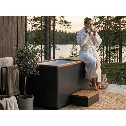 Frau im weißen Bademantel nutzt Swedish Cold Generation 4S Eisbad mit schwarzem Design auf einer Holzterrasse mit Seeblick und Waldkulisse