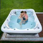 Wellis Lugano Life Premium-Whirlpool mit zwei Personen im Garten, sprudelnde Düsen und blaues Wasser sichtbar