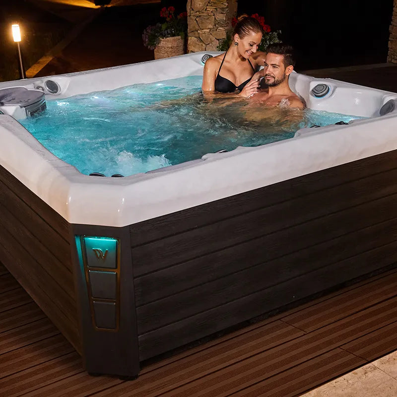 Wellis Palermo Life Premium-Whirlpool mit zwei Personen im türkisblauen Wasser auf Terrasse bei Nacht