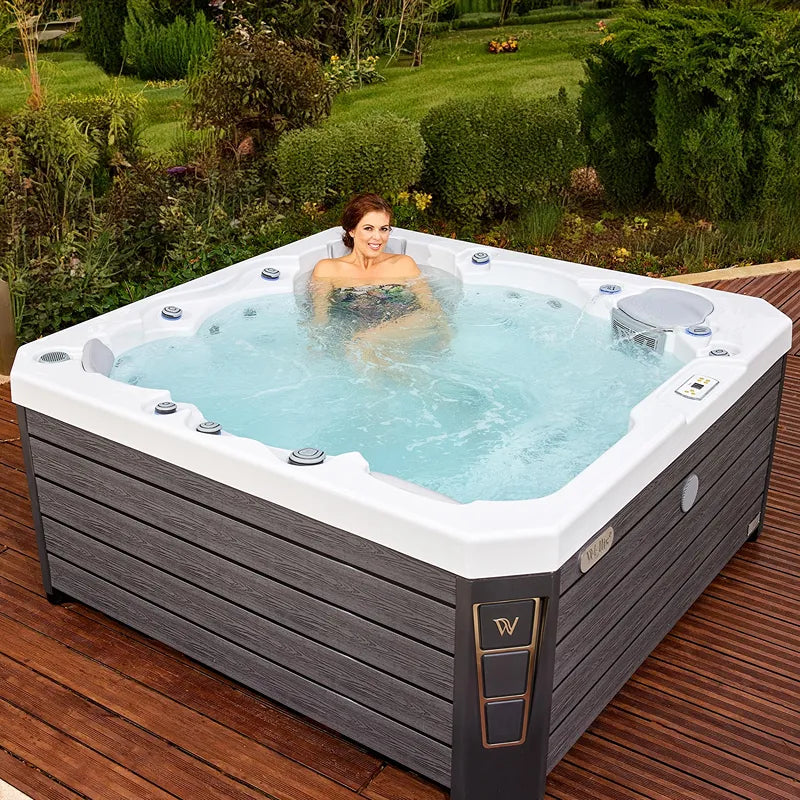 Wellis Prague Life Premium hot tub with woman in turquoise blue water, white tubs and dark grey wooden cladding in garden